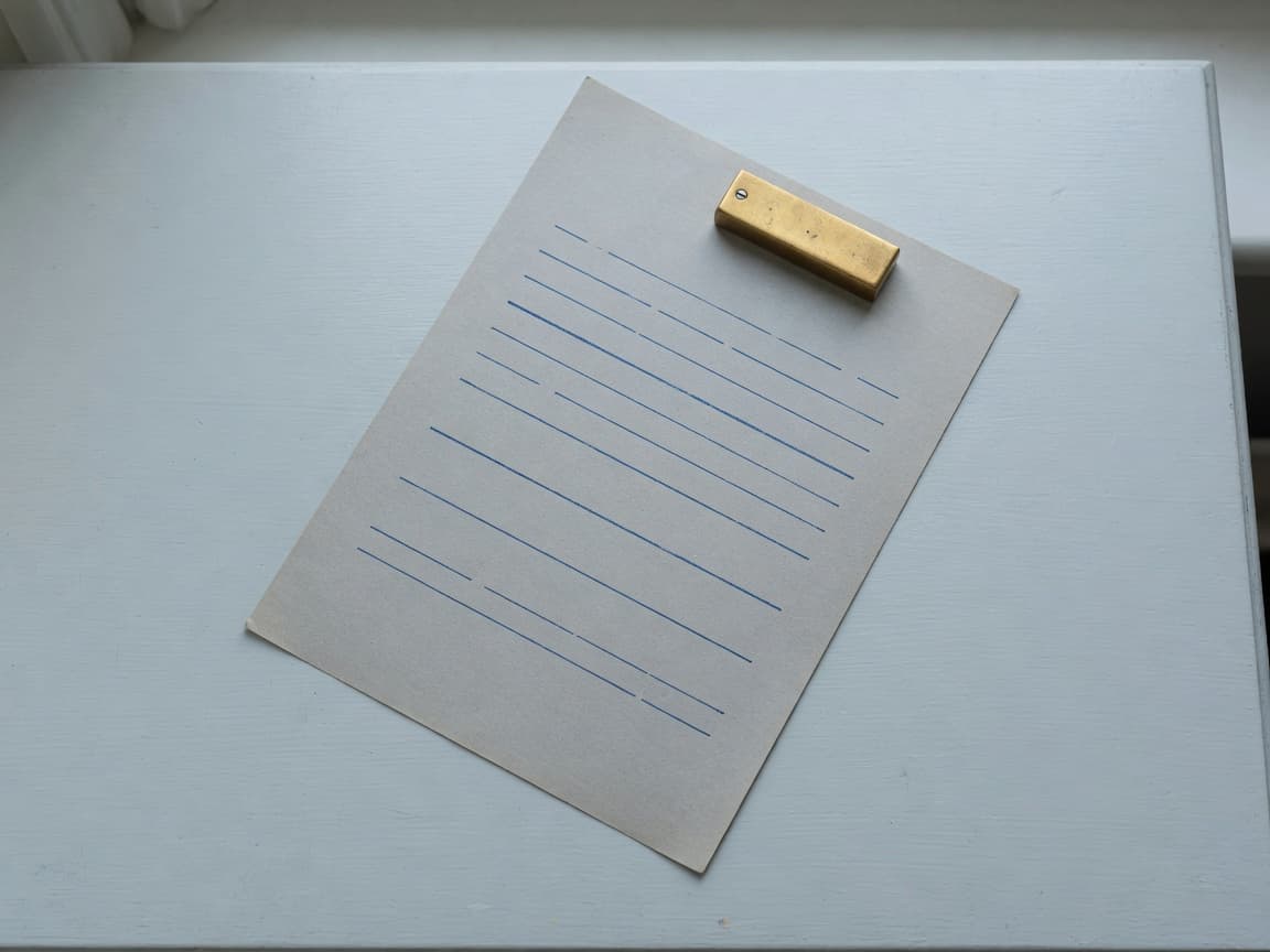 A sheet of ruled paper with cobalt lines and a brass weight on a pale desk.